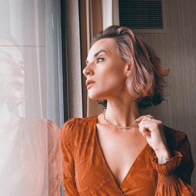 Brooks leaning against and staring out a window while wearing a burnt-orange dress