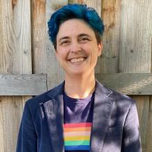 a middle-aged white person with blue hair smiling outdoors, in a blazer and rainbow T-shirt